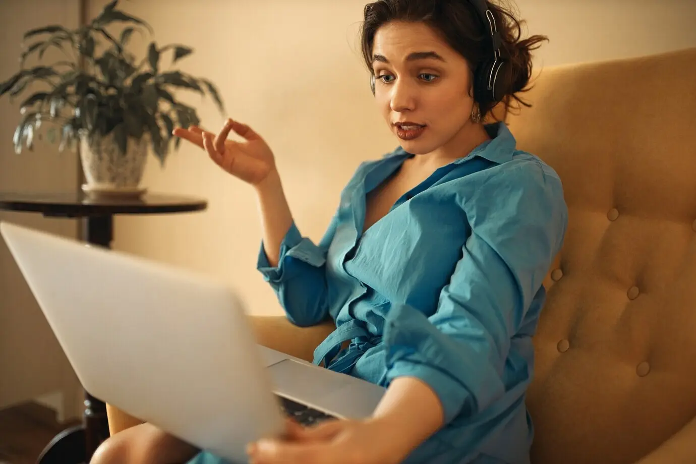 A stylish, confident young woman wearing headphones sits on a sofa with a laptop, participating in a group business online meeting via a webcam conference chat, gesturing emotionally while brainstorming with colleagues.
