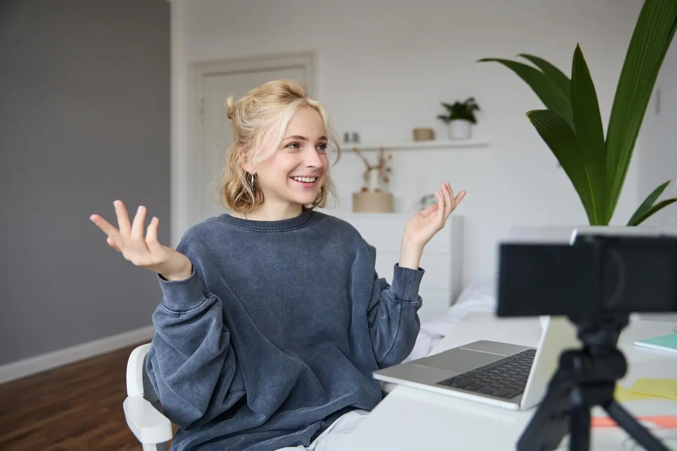 A portrait of a young woman who creates social media content, sitting in a room with a laptop and a digital camera.