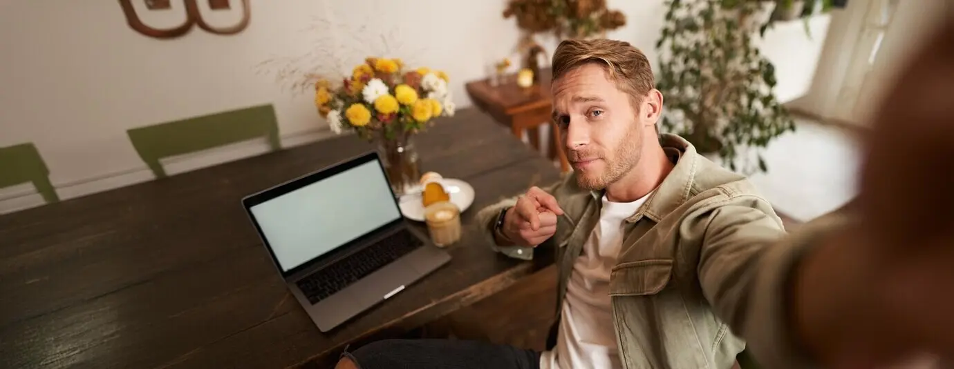 A handsome, confident young man, seated in a cafe with a laptop and working from the coffee shop, takes a selfie on his mobile phone, pointing his finger at the camera with a sassy expression.